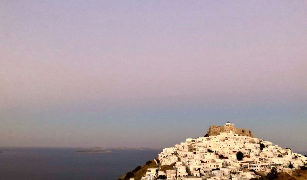 Whitewashed houses cascade down the hillside of Astypalaia, Greece, toward the sea, crowned by a historic hilltop castle at sunset.