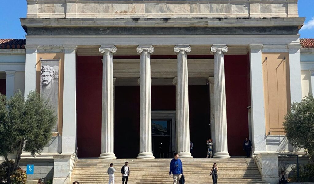 Front entrance of the National Archaeological Museum in Athens, Greece, featuring tall marble columns, a red facade, and visitors walking on the steps.