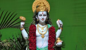 A statue of a Hindu deity adorned with a golden crown, jewelry, and a garland of red flowers, holding a golden discus and a small pot, set against a bright green background with palm leaves.