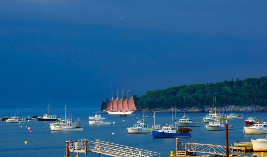 A colorful schooner with red sails glides through the harbor at Bar Harbor, Maine, surrounded by fishing boats and backed by a forested shoreline.