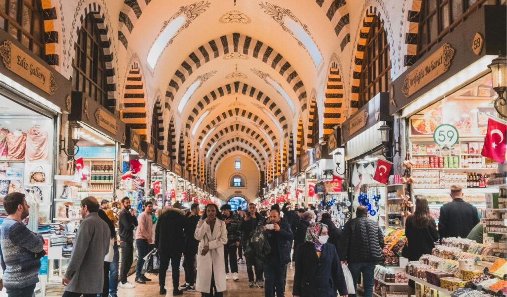 Crowds walk through a colorful indoor bazaar lined with market stalls, showcasing spices, textiles, and souvenirs.