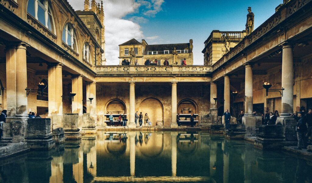 Historic Roman Baths in Bath, England, with columns, statues, and reflections in the green thermal pool surrounded by visitors.