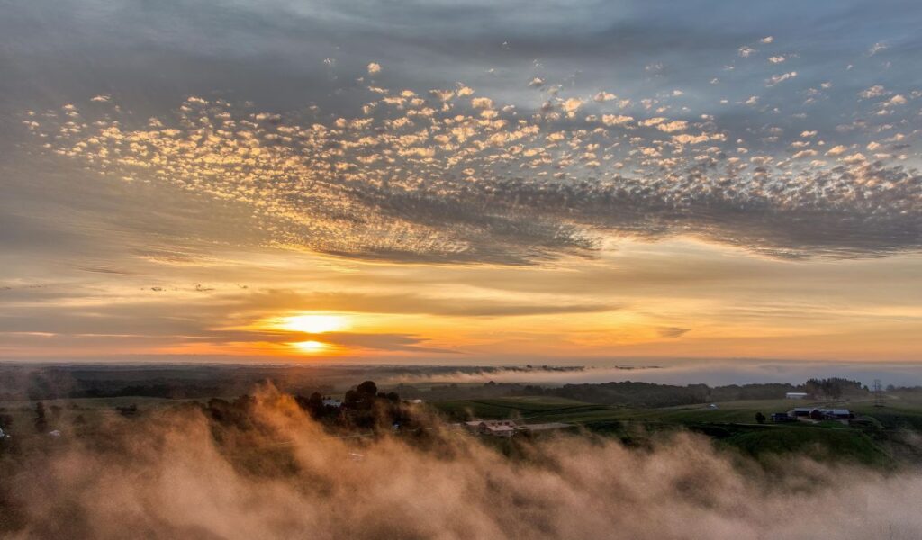Golden sunrise over Bayfield, Wisconsin, with mist rising above rolling green farmland and a dramatic, cloud-streaked sky.