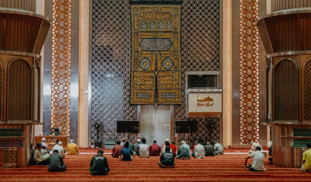 People sitting and praying inside a beautifully decorated mosque with intricate Islamic patterns and a serene atmosphere.