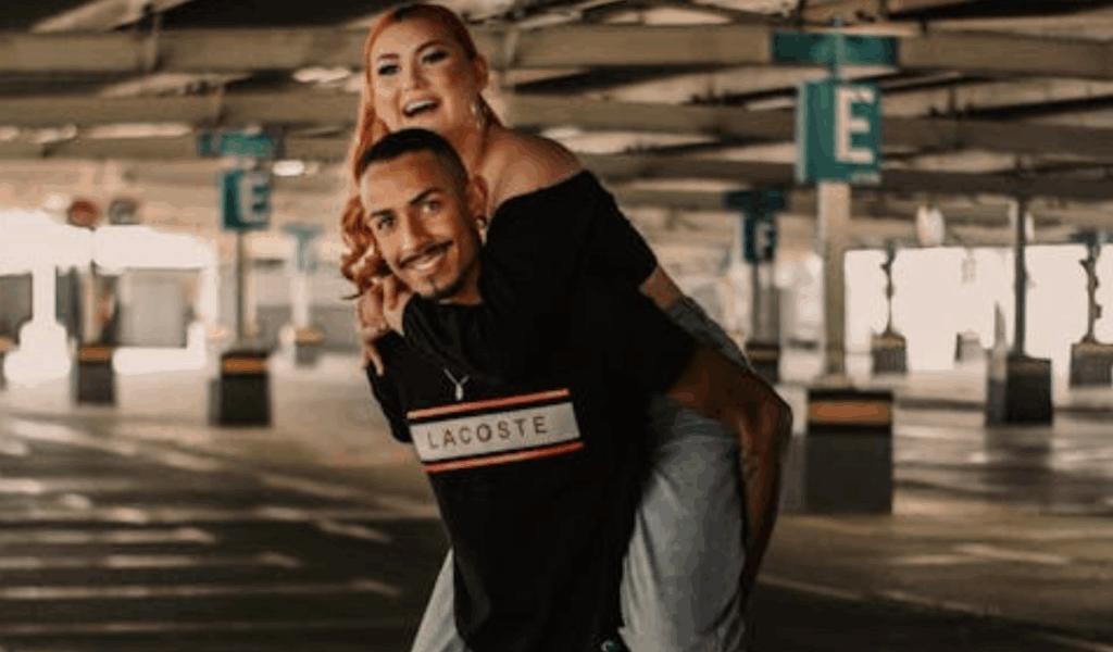 A smiling man gives a piggyback ride to a laughing woman in a parking garage, capturing a playful and affectionate moment between them.