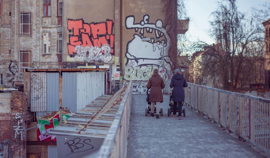 Two people pushing strollers walk across a graffiti-covered pedestrian bridge in Berlin, with colorful street art and old buildings in the background.