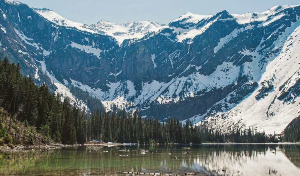 Snow-capped mountains towering over a clear alpine lake in Bigfork, Montana, surrounded by dense evergreen forests and blue skies.
