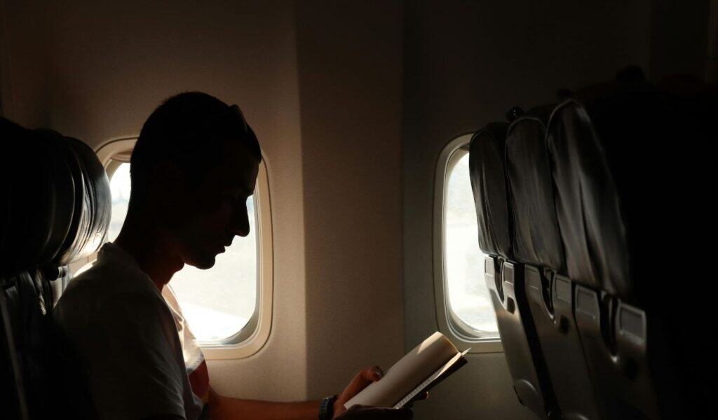 Silhouette of a man sitting by an airplane window reading a book, with sunlight streaming in and empty seats beside him.