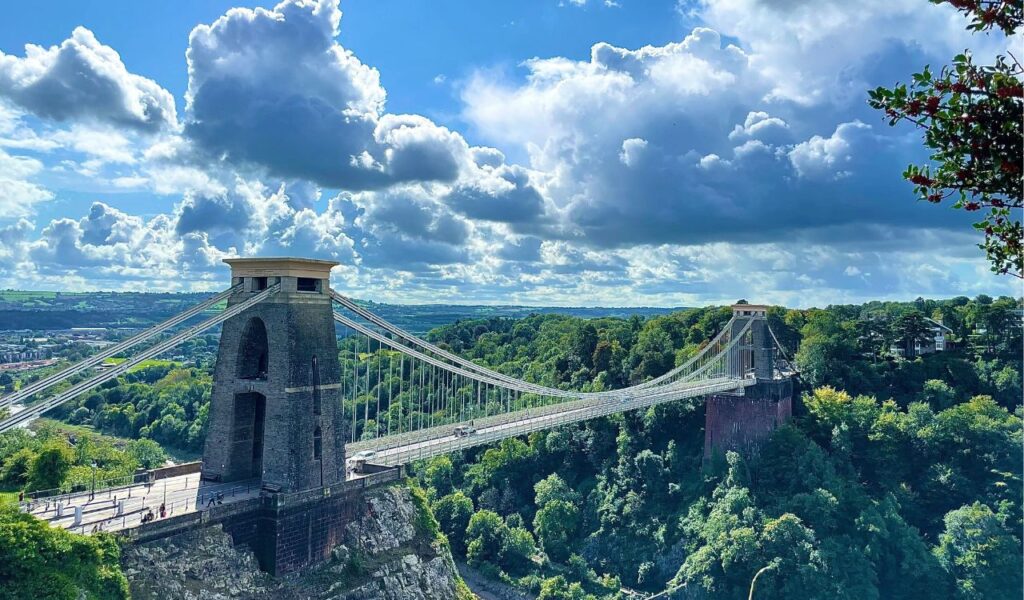  Clifton Suspension Bridge in Bristol spanning the Avon Gorge, surrounded by lush green hills under a partly cloudy sky.








