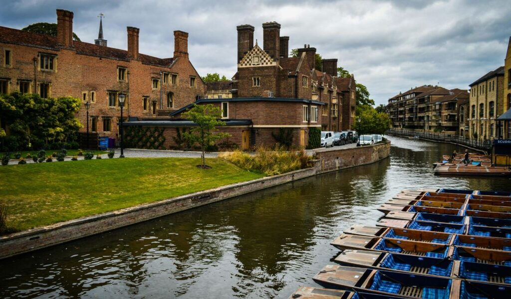 Wooden punts lined along the River Cam in Cambridge, with historic brick college buildings and a green lawn on the riverbank.