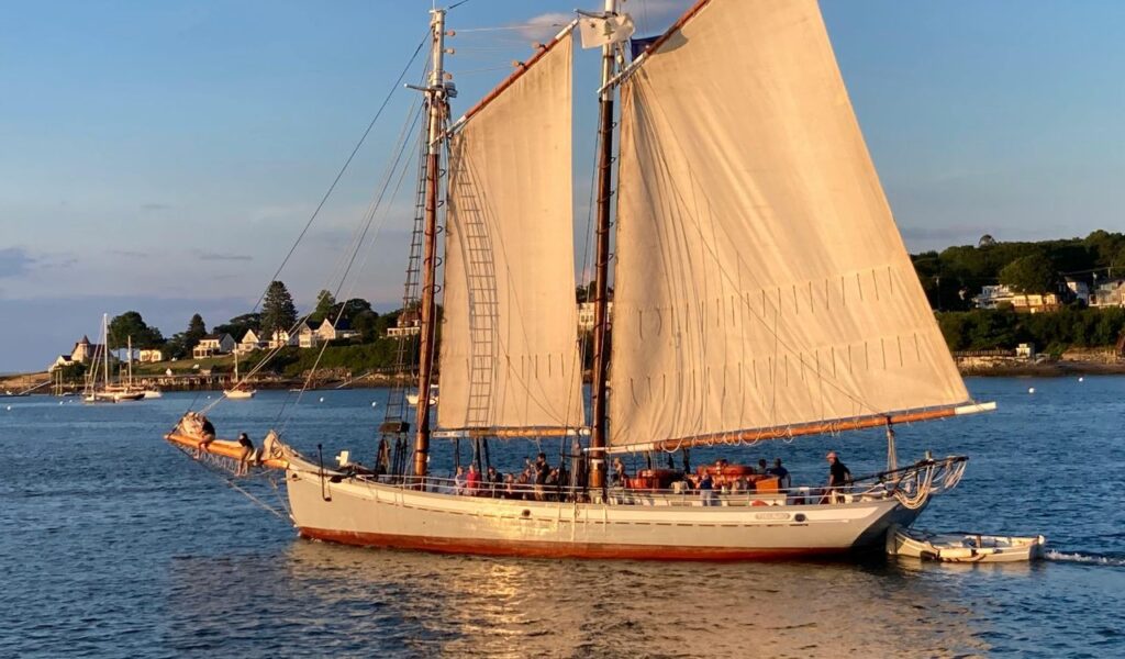 A classic sailboat glides through Camden’s harbor at sunset, with passengers on board and coastal homes in the background.
