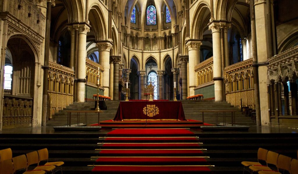Ornate interior of Canterbury Cathedral with tall stone arches, stained glass windows, and a red-carpeted altar at the center.