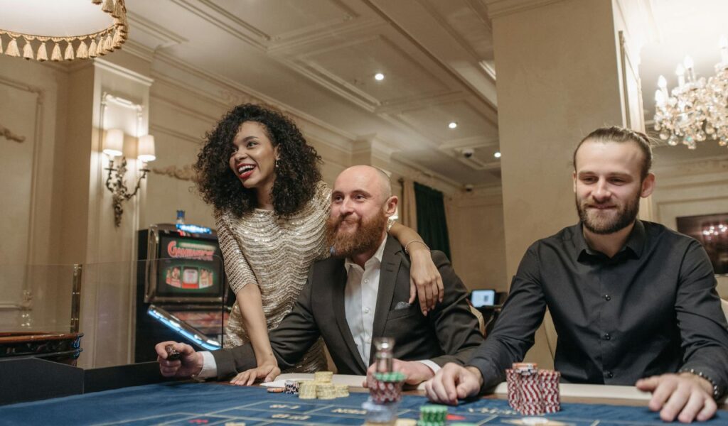 Three people enjoy a casino game at a table, smiling and surrounded by poker chips in an elegant room with chandeliers and slot machines.