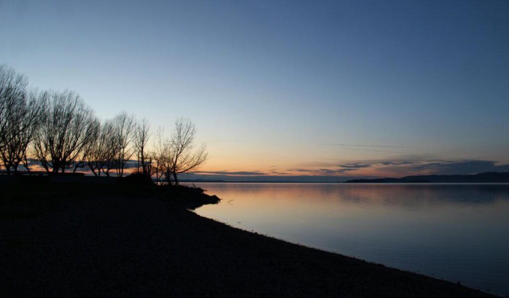 Peaceful sunset over Lake Trasimeno in Castiglione del Lago, Italy, with silhouetted trees along the shore and calm, reflective waters.