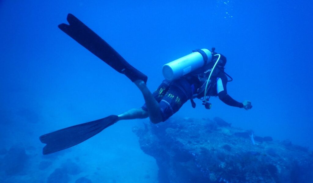 A scuba diver swims underwater in deep blue ocean water, equipped with diving gear and fins, exploring near a rocky coral-covered area.