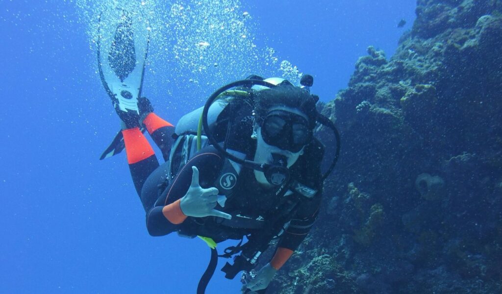 A smiling scuba diver underwater gives a hand gesture while wearing full diving gear, with bubbles rising around and rocky coral formations nearby