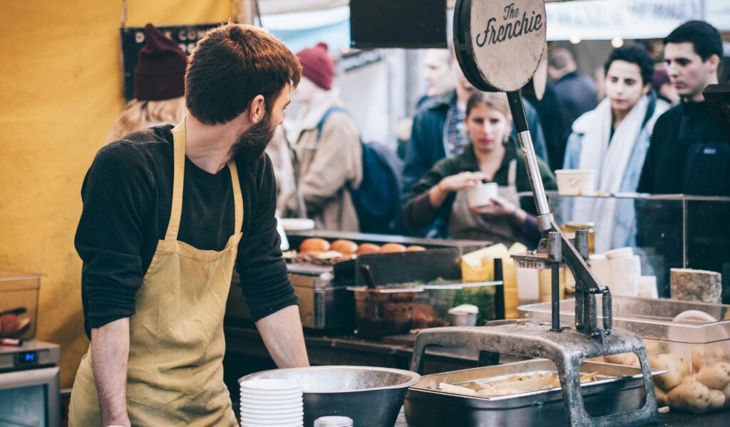 A man wearing a yellow apron works at a busy food stall, preparing meals as customers wait in line at an outdoor market.