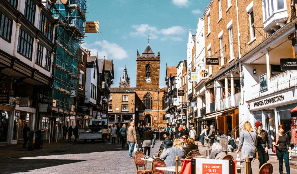 A lively street scene in Chester, Connecticut, with people dining outdoors, shopping, and strolling past historic brick buildings under a bright blue sky.