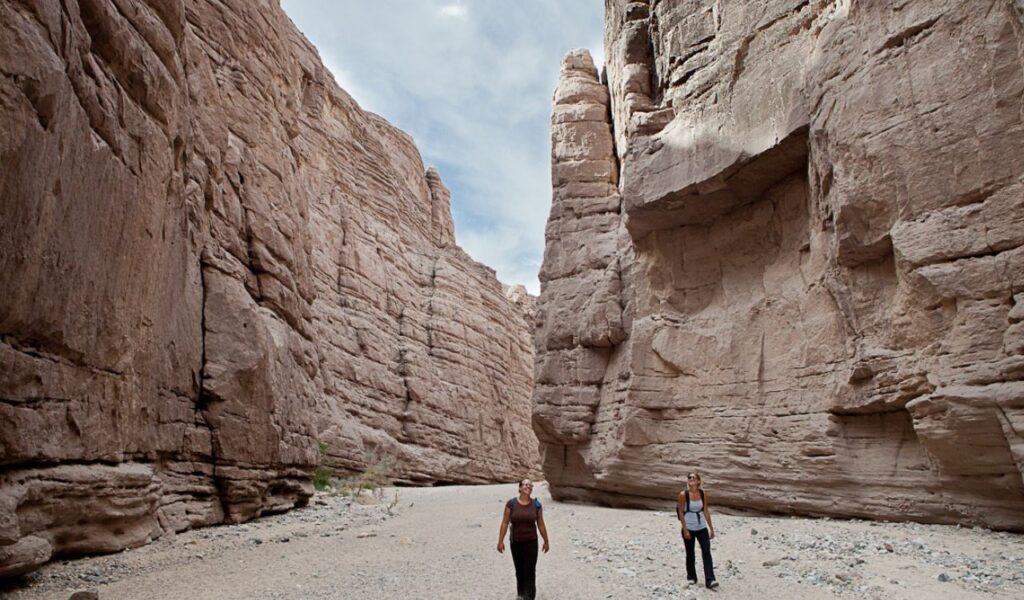 Two hikers walk through a wide slot canyon with towering beige rock walls at Chuckwalla National Monument in California under a partly cloudy sky.