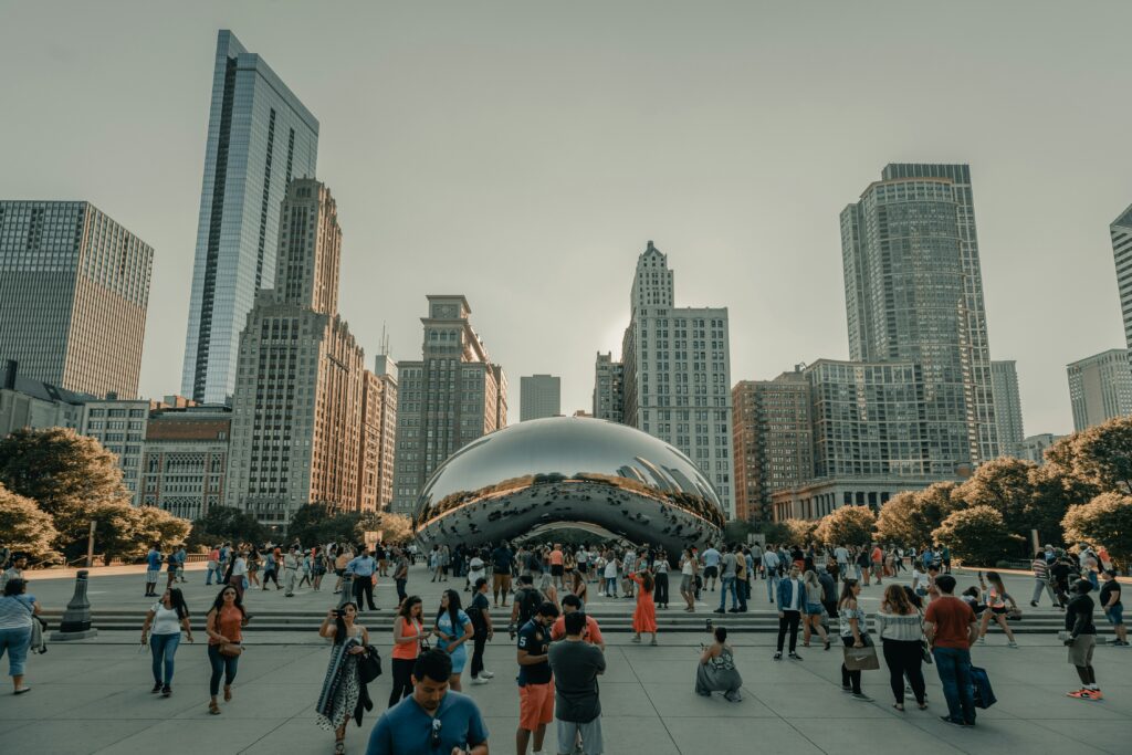 A large crowd of tourists gathers around the reflective Cloud Gate sculpture, also known as "The Bean," in Millennium Park, Chicago, surrounded by tall skyscrapers under a clear sky.