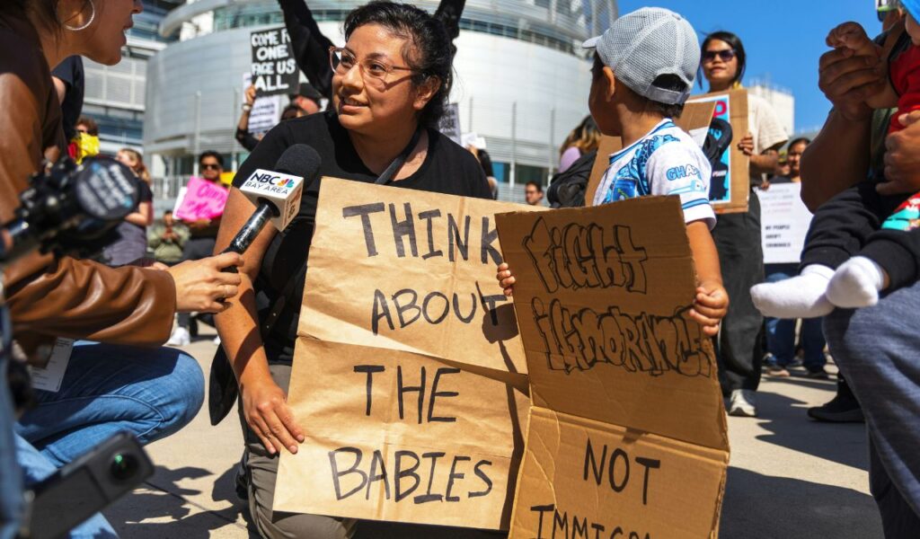 A woman holding protest signs reading 'Think About the Babies' and 'Fight Ignorance Not Immigrants' speaks to a reporter during a political rally.