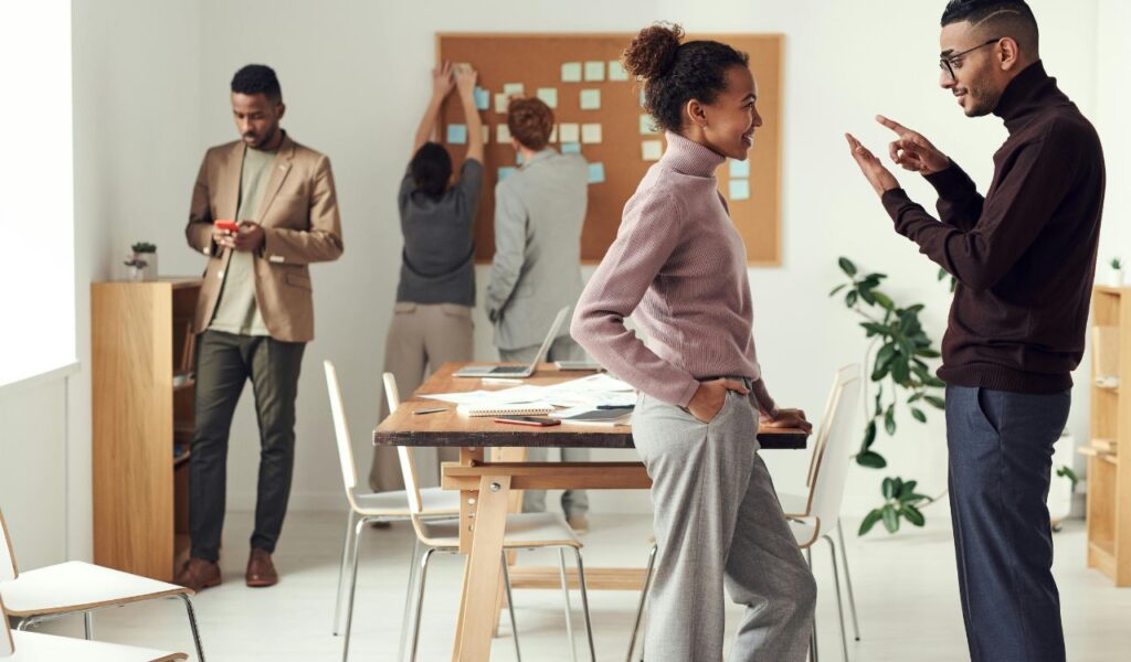 Group of colleagues in an office, with two people having a focused conversation while others work on a corkboard in the background.