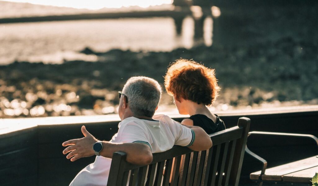 An older couple sitting closely on a wooden bench, facing the ocean and enjoying a peaceful conversation during sunset.