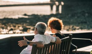 An older couple sitting closely on a wooden bench, facing the ocean and enjoying a peaceful conversation during sunset.