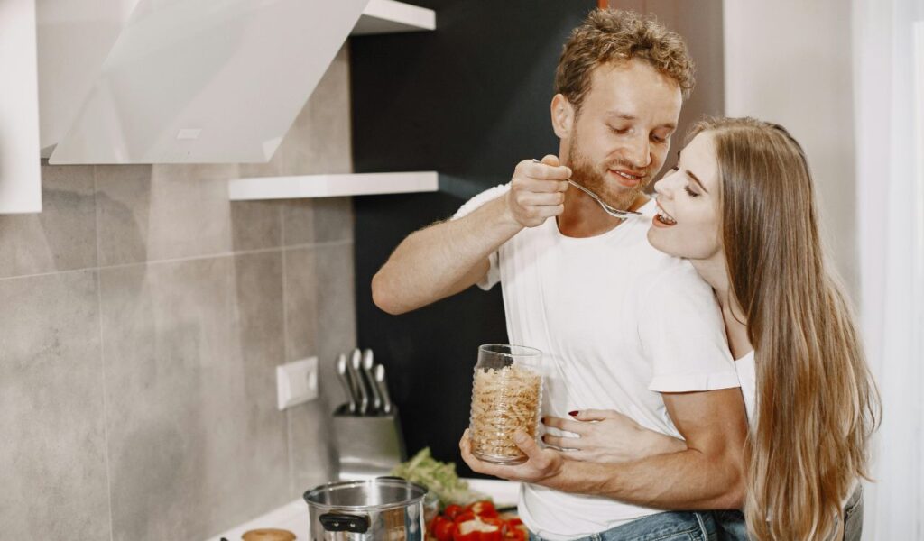 A smiling couple enjoys cooking together in a modern kitchen, with the man feeding the woman a taste from a spoon while she hugs him from behind.