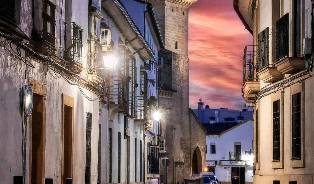 Narrow cobblestone street in Córdoba, Spain, lined with whitewashed buildings and wrought-iron balconies, leading to a stone archway at sunset.