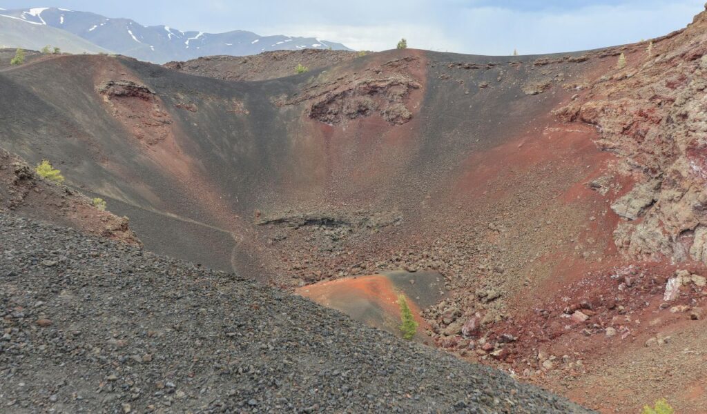 Volcanic crater at Craters of the Moon National Monument in Idaho, with dark lava rock, red earth tones, and sparse vegetation.