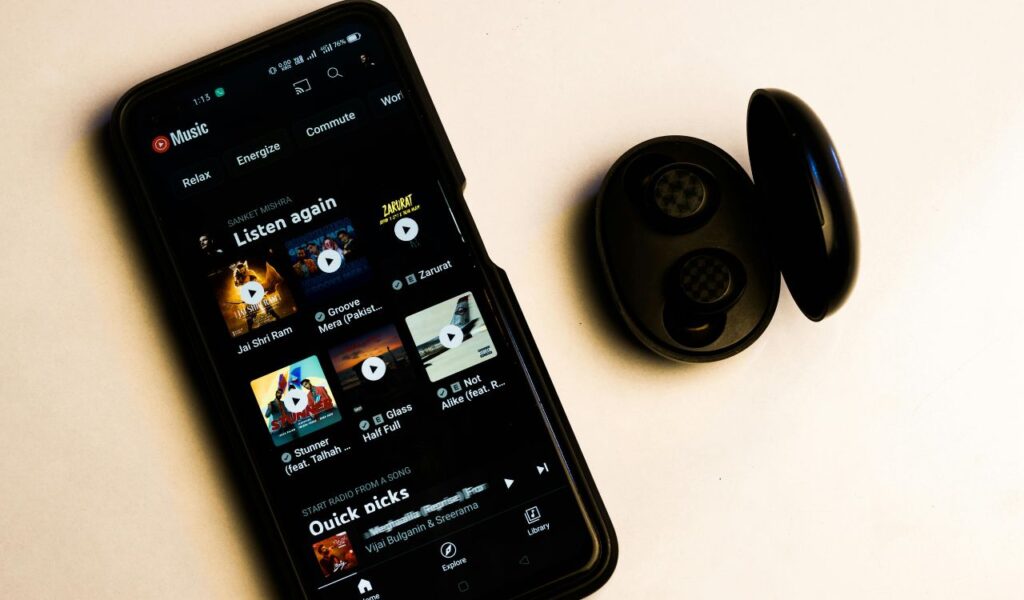 A smartphone displaying a music streaming app with playlists and song recommendations, placed next to an open case containing wireless earbuds.