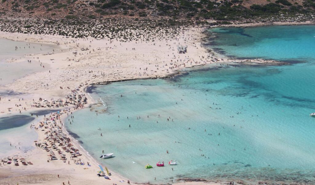 Aerial view of Balos Beach in Crete, Greece, with its white sand, shallow turquoise waters, and people relaxing along the shore.