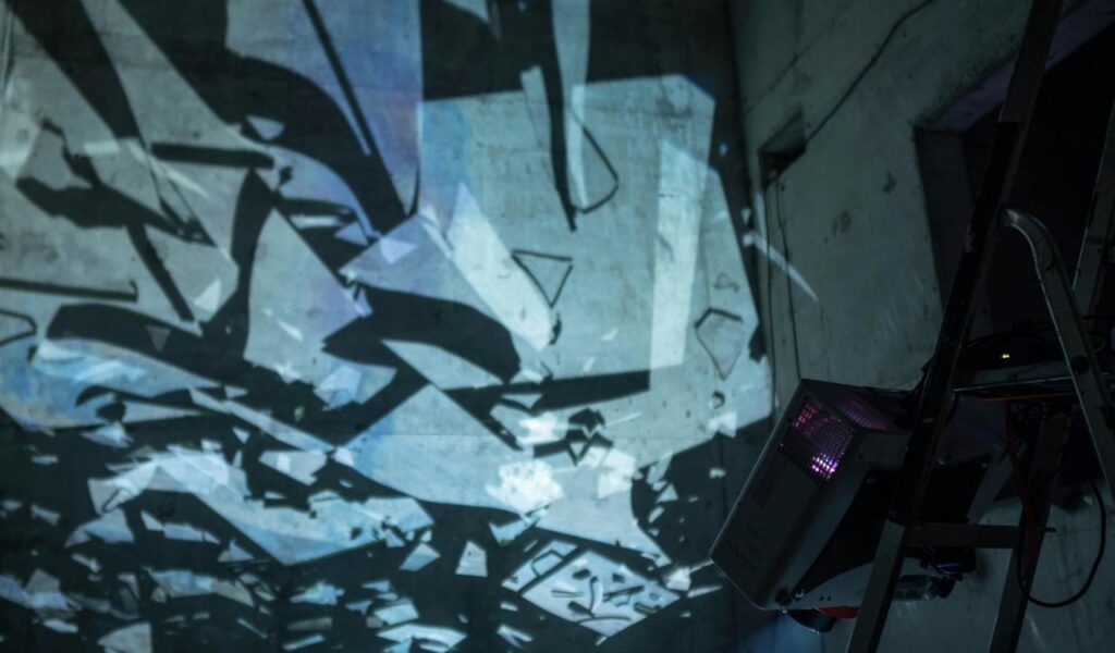 A wall with a projection of shattered glass patterns illuminated in blue tones, creating an abstract effect, with a projector visible on the side.