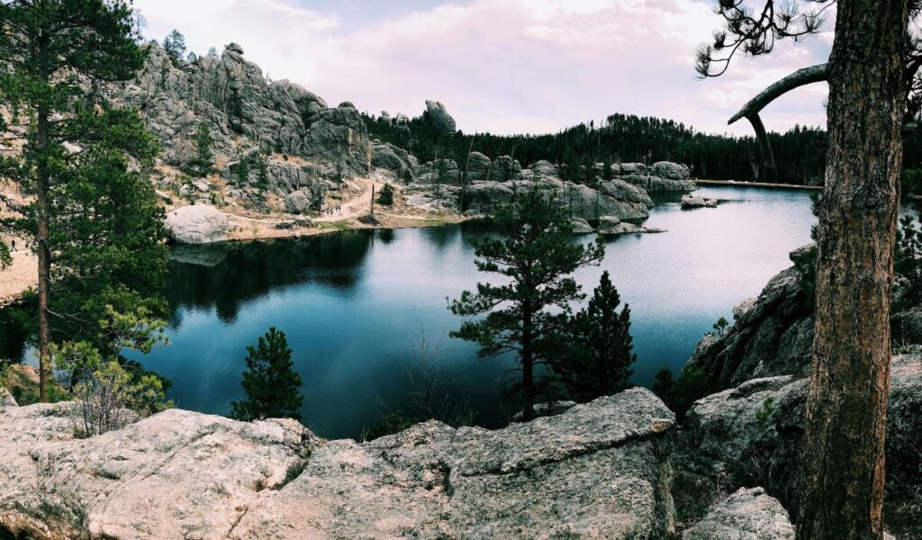 A serene lake surrounded by rocky cliffs and pine trees under a partly cloudy sky in Deadwood, South Dakota.