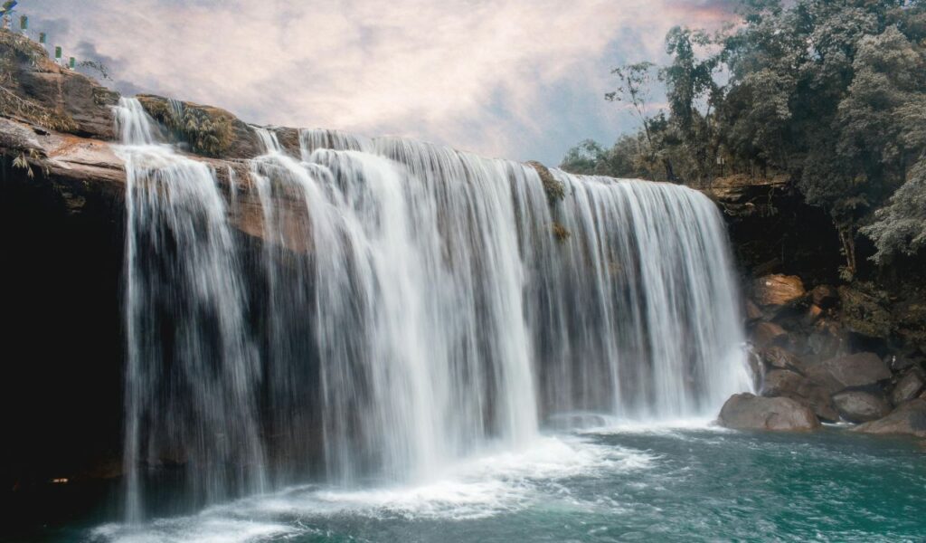 Wide curtain of water at DeSoto Falls cascading over a rocky ledge into a turquoise pool, framed by lush green forest and rocks.