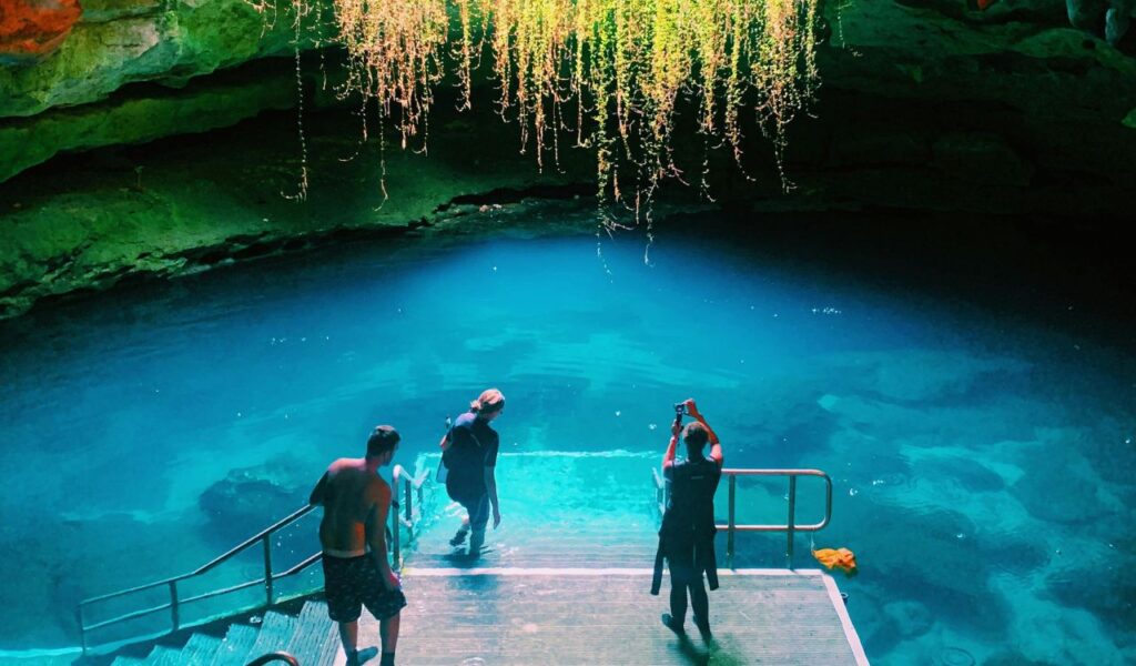 People descending steps into the glowing blue waters of Devil’s Den cave in Florida, surrounded by rocky walls and hanging greenery.