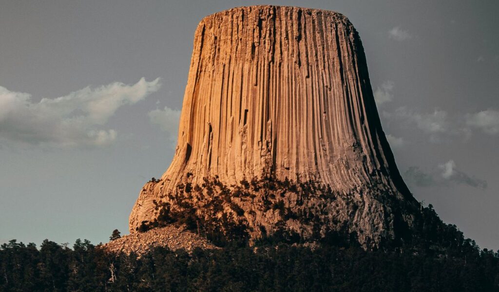 Towering monolithic rock formation of Devils Tower in Wyoming, rising steeply above surrounding forest under a partly cloudy sky.