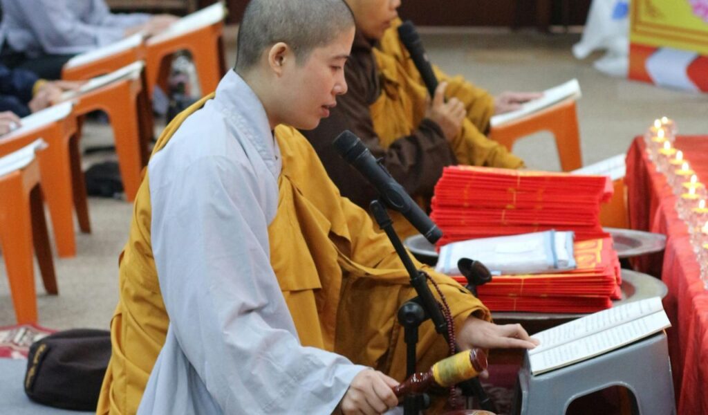 Buddhist monks in traditional robes chant and read scriptures inside a temple, with microphones and stacks of red prayer books on a table beside them.