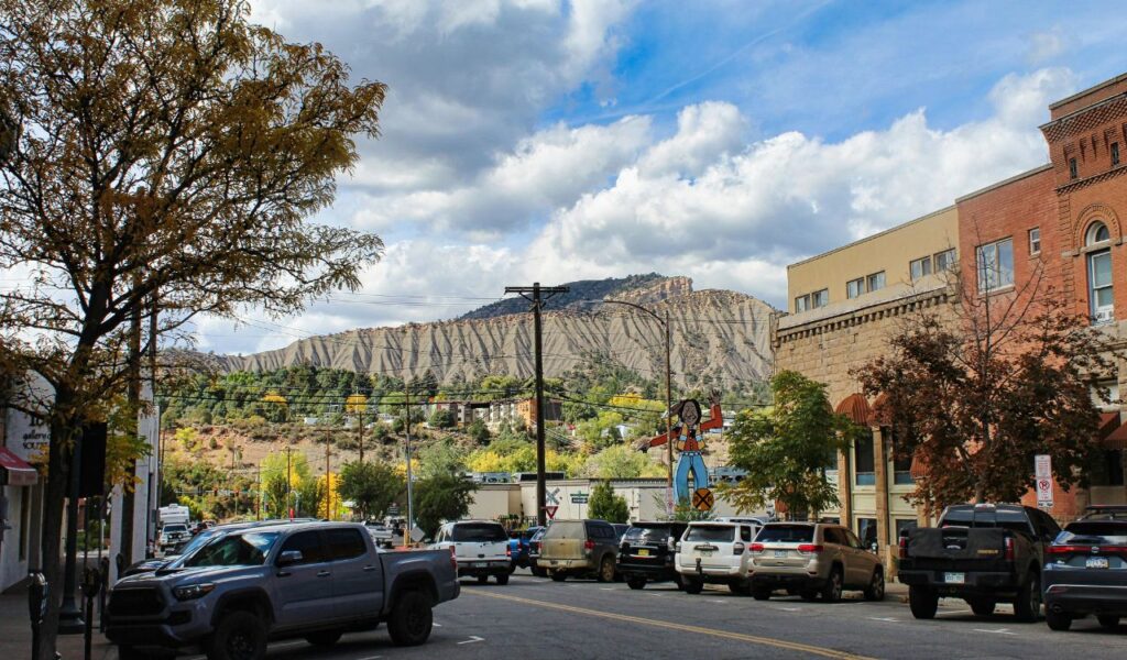 Street view in Durango, Colorado, with parked cars, brick buildings, trees, and rugged mountains in the background under a partly cloudy sky.
