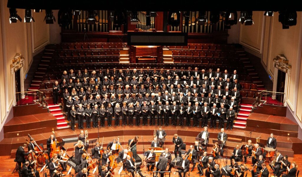 Orchestra and large choir performing in a grand Edinburgh concert hall with elegant seating, red carpets, and organ pipes.