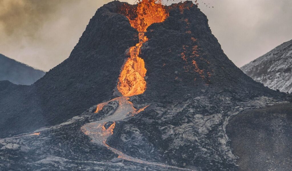 A volcano at El Malpais National Monument in New Mexico erupts, sending bright orange lava cascading down its dark rocky slopes.