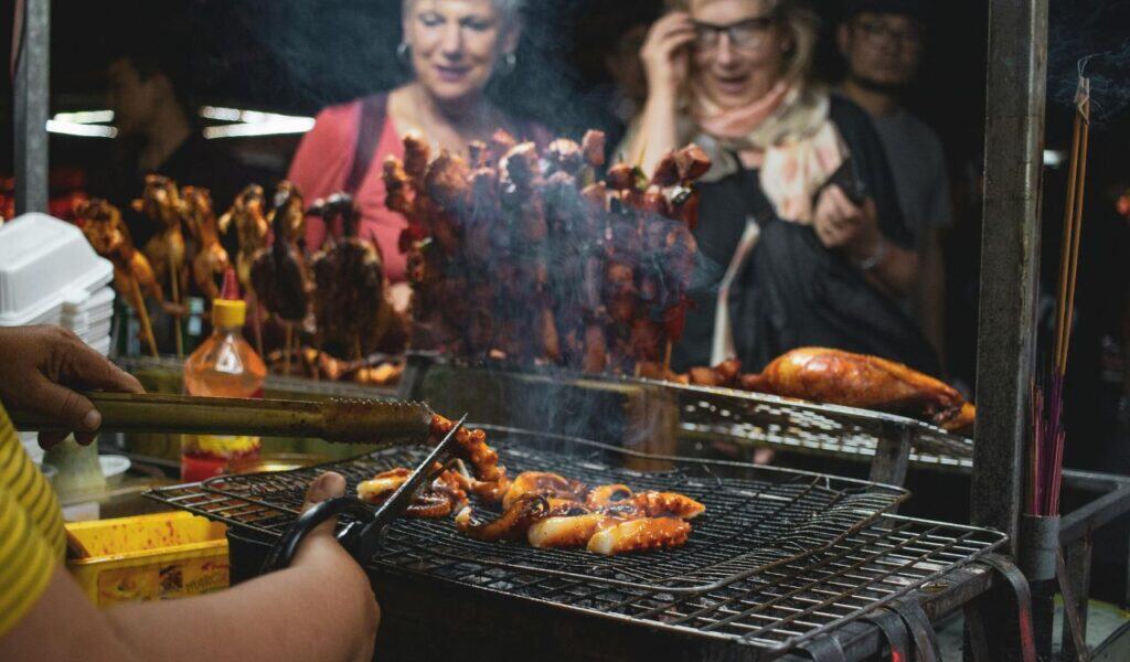 Close-up of a street food vendor grilling octopus tentacles, with skewered meats in the background and curious customers watching.