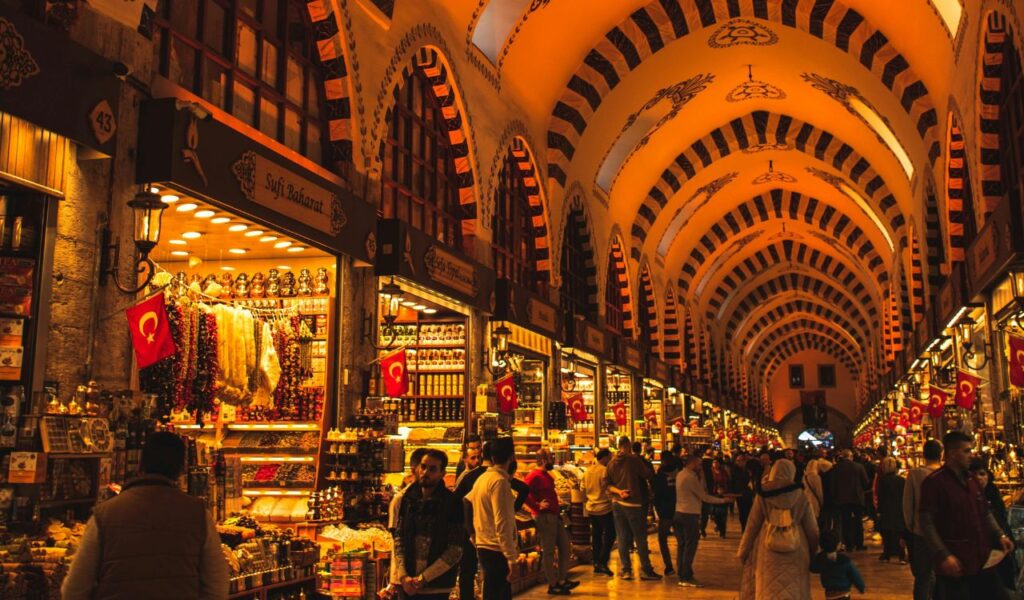 Tourists and locals interact inside a vibrant Middle Eastern market lined with spice shops and traditional goods.