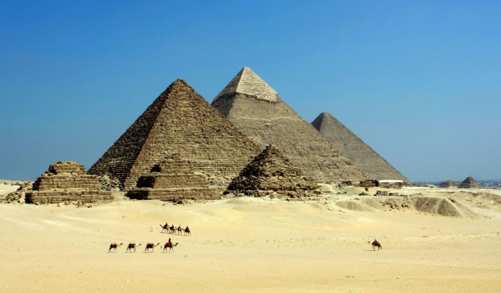 Distant view of Egyptian pyramids in the desert with a group of camels passing in front.
