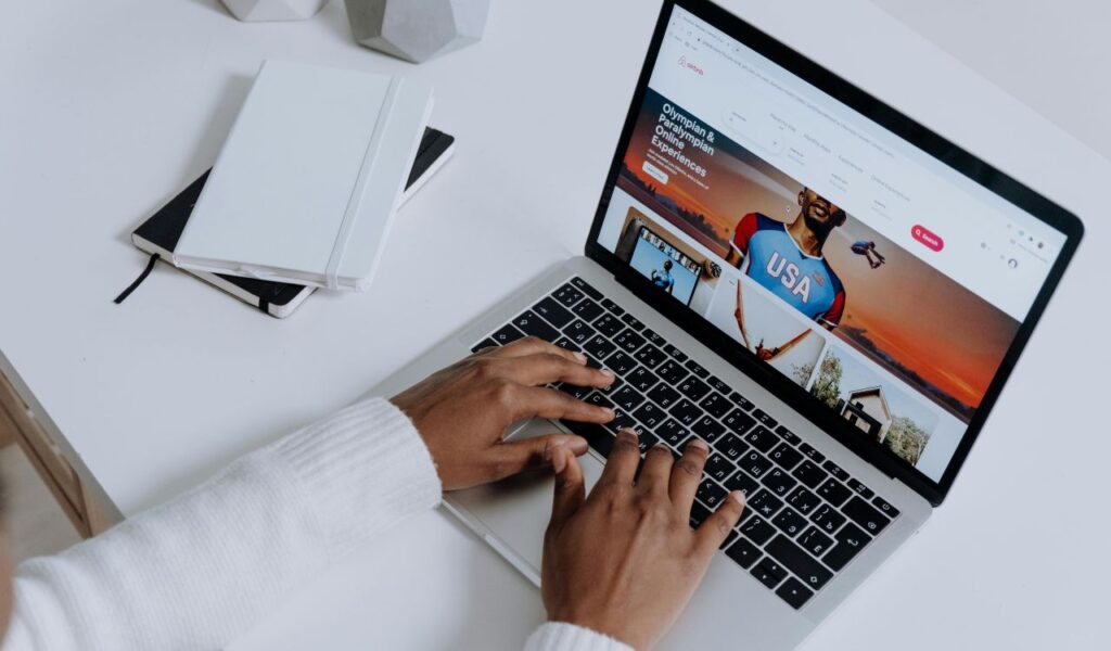 A person uses a laptop displaying the Airbnb website, with hands on the keyboard and notebooks placed on a white desk nearby.