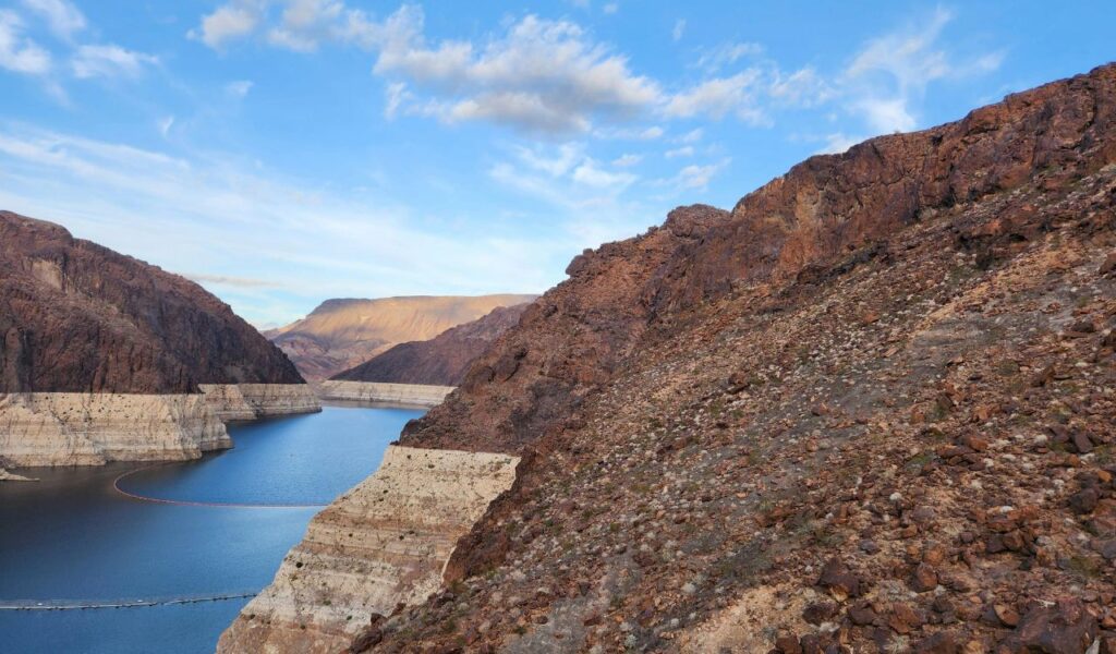 Rocky landscape surrounding Hoover Dam with low water levels visible in the reservoir under a blue sky.