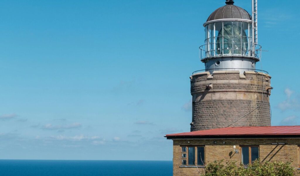 Close-up view of the historic Fire Island Lighthouse in New York, with its stone tower, red-roofed building, and blue ocean backdrop.