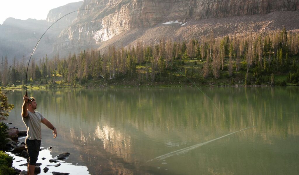 A man casts a fishing line into a calm, reflective lake surrounded by pine trees and rocky cliffs at First Landing State Park in Virginia.