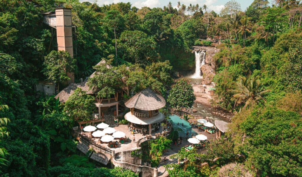 An aerial view of a tropical riverside resort in Bali, surrounded by lush jungle, with thatched-roof buildings, white sun umbrellas, a swimming pool, and a scenic waterfall cascading into the river in the background.